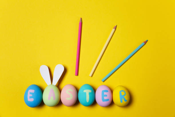 istock Row of colorful pastel monophonic painted Easter eggs with inscription Easter, fun bunny ears on egg, three pencils on yellow background. Copy space for advertisement. With place for text. Top view. 908398558