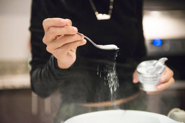 This is a color, royalty free photograph of a young 20 year old Millennial woman baking cupcakes from scratch at home in the kitchen in Orlando, Florida, USA. She measures out a spoon full of baking soda to add to the bowl of batter.