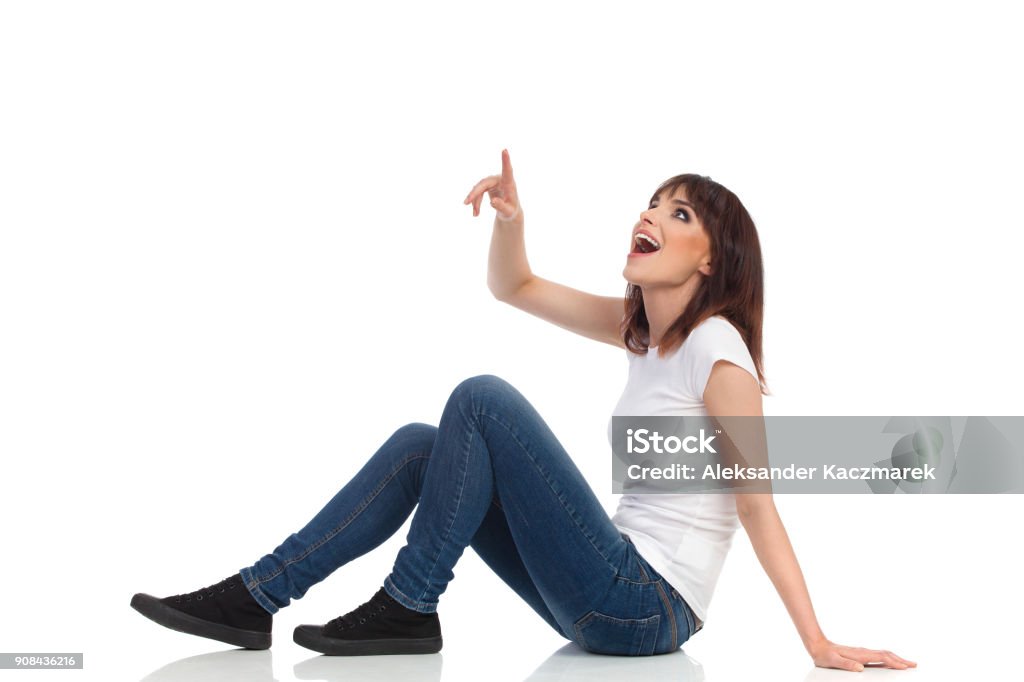 Excitada joven está sentado en el piso, señalando, mirando y gritando. Vista lateral - Foto de stock de Sentado en el suelo libre de derechos