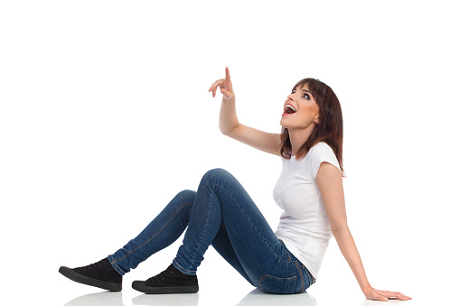 Young woman is sitting on a floor, pointing, looking up and shouting. Side view. Full length studio shot isolated on white.