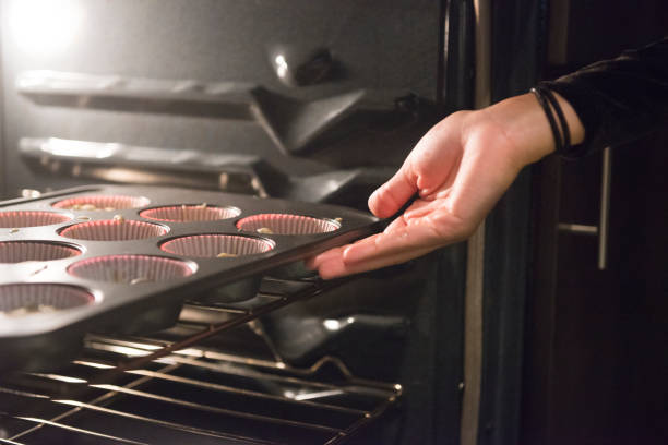 This is a color, royalty free photograph of a young 20 year old Millennial woman baking cupcakes from scratch at home in the kitchen in Orlando, Florida, USA. She places the tin on a shelf in the oven for baking.