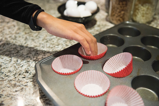 This is a close up color, royalty free photograph of a young 20 year old Millennial woman baking cupcakes from scratch at home in the kitchen in Orlando, Florida, USA. She places the empty wrappers in a metal baking tin.