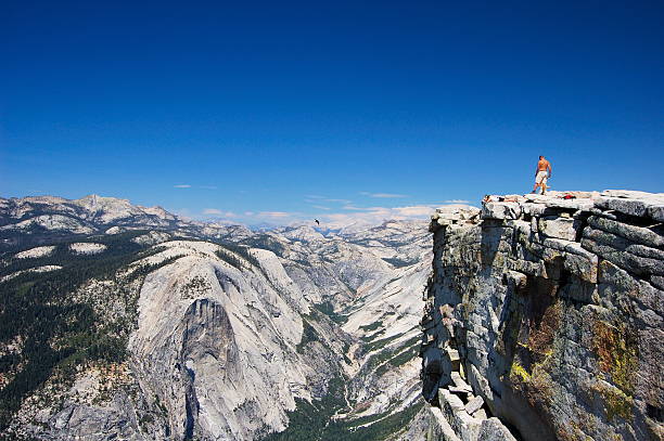 This stunning photograph captures the iconic Half Dome and surrounding granite cliffs in Yosemite National Park, California. A winding path leads through a peaceful forest of tall trees, drawing the viewer's eye toward the breathtaking rock formations in the distance. The blue sky dotted with wispy clouds adds to the serene and awe-inspiring atmosphere of this natural wonder.