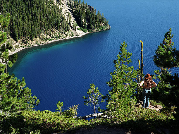 A girl in a brown cowboy hat with long brown hair and blue jeans looking over the very blue Crater Lake in Oregon surrounded by evergreen pine trees. With hands on hips, she is taking in the awesome beauty of nature on a gorgeous sunny day.