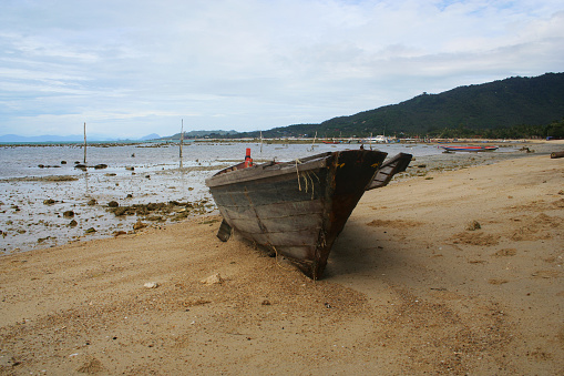 The boat of a fisherman on the beach of Koh Samui (Thailand) during ebbs is on the sand of the beach. In the background some parts of the island and the cloudy sky. Extreme perspective shot from the front of the tiny boat.