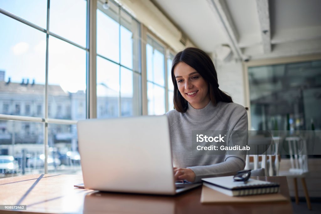 Smiling attractive female freelancer doing remote job using laptop computer Connected to wireless internet in office, professional administrative manager checking online business report documentation People Stock Photo