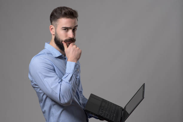 istock Confused young business man using laptop computer touching beard and looking at camera 908605184