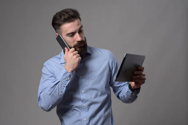 istock Busy young smart casual bearded man talking on the mobile phone and watching at tablet computer 908605492
