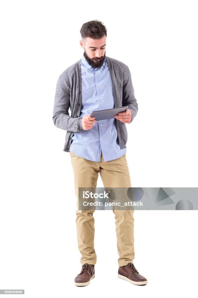 Young adult bearded hipster holding and watching at tablet computer. Young adult bearded hipster holding and watching at tablet computer. Full body length portrait isolated on white studio background. Men Stock Photo