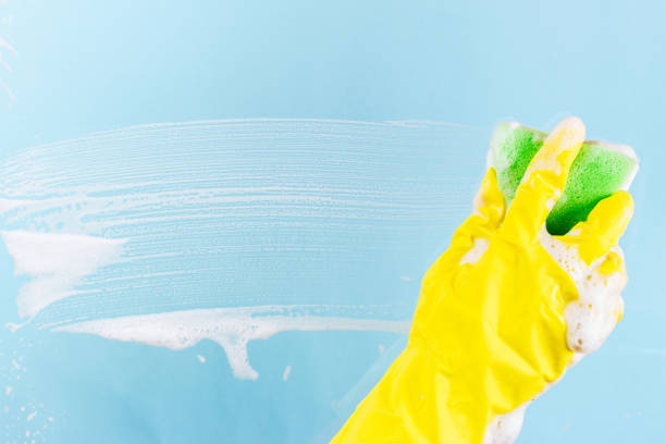 istock Washing windows and glass. The worker in a yellow glove can stack a sponge with foam on a blue background 908617202