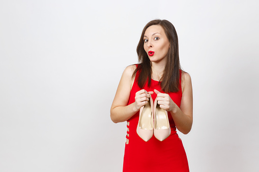 Smiling glamour caucasian fashionable young smiling brown-hair woman in red dress holding pair of beige shoes with red sole of her shopping isolated on white background. Copy space for advertisement