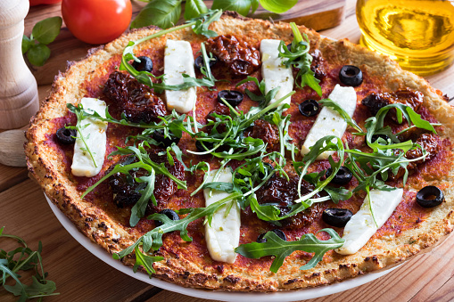 Gluten-free grain-free vegetarian pizza made from mashed cauliflower and almond flour, topped with sundried tomatoes, olives, goat cheese and fresh arugula.