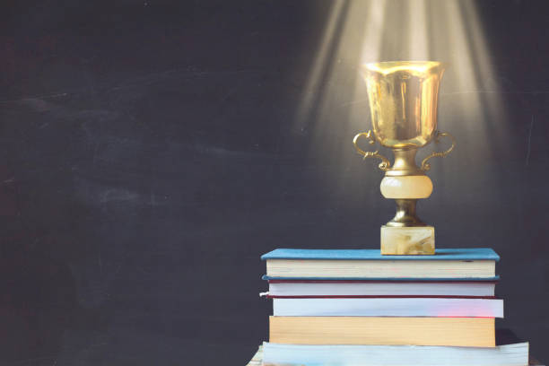 trofeo de oro en la pila de libros, contra la pizarra, con rayos de sol sobre trofeo; concepto de aprendizaje/logros - honra fotografías e imágenes de stock