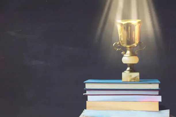 Photo of Golden trophy on pile of books, against blackboard, with sun rays over trophy; learning/achievement concept