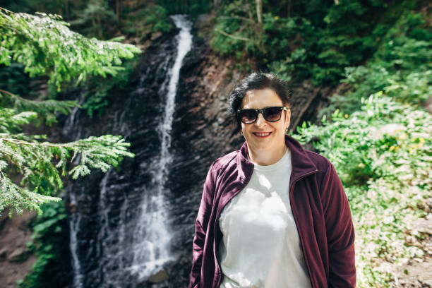 istock Middle-aged woman in sunglasses posing against waterfall background 908749828