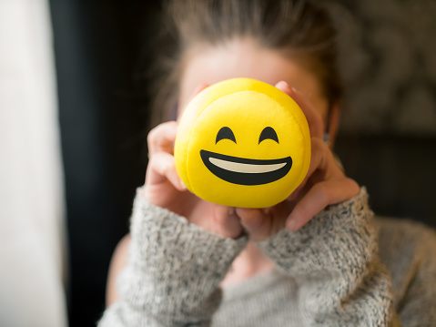 Girl holding smiley face laughing emoticon in front of her face. The young woman is of caucasian ethnicity and is casually dressed. The scene is situated indoors in a city apartment in Sofia, Bulgaria (Eastern Europe) during the day. The footage was taken with Panasonic GH5 camera.