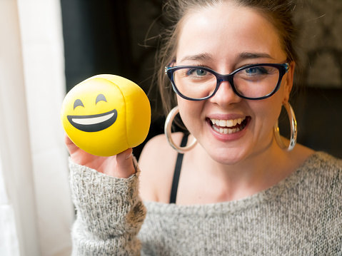 Girl holding emoticon smiley face and laughing. The young woman is of caucasian ethnicity and is casually dressed. The scene is situated indoors in a city apartment in Sofia, Bulgaria (Eastern Europe) during the day. The footage was taken with Panasonic GH5 camera.