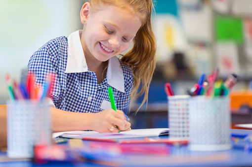 Schoolgirl working at a desk. She is wearing a school uniform. She is writing or drawing with a felt pen. She is smiling and happy having fun.
