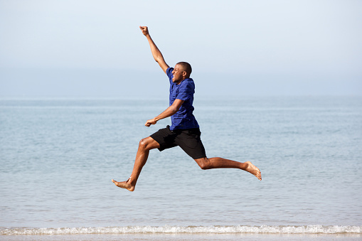 Full body portrait of excited young african guy running and jumping along the sea