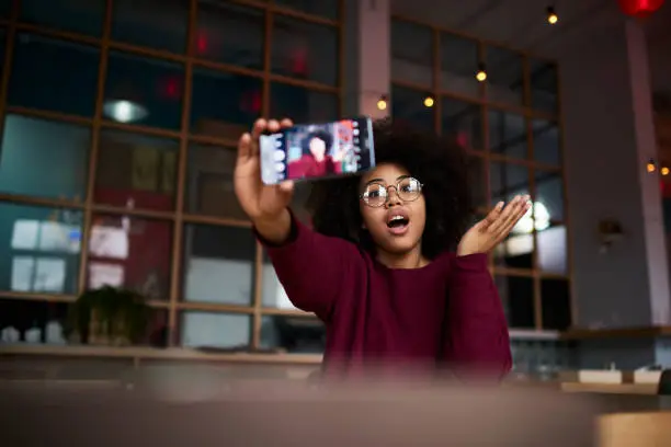 Emotional young afro american girl with curly hair in trendy glasses posing while making photo for updating account picture using modern smartphone camera and wifi access to internet in cafe indoors
