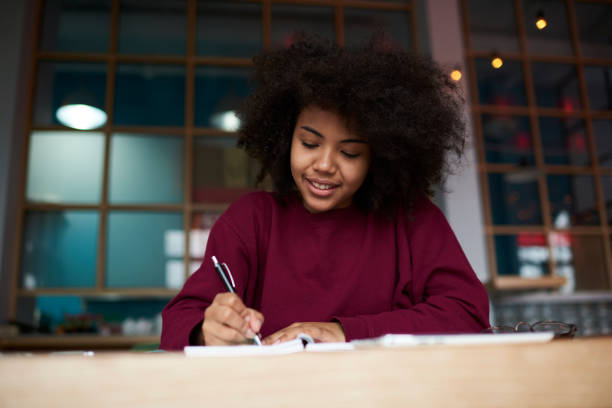 istock Positive concentrated afro american female student preparing homework task writing composition on literature sitting in university library, talented journalist creating article noting ideas with pen 908881170