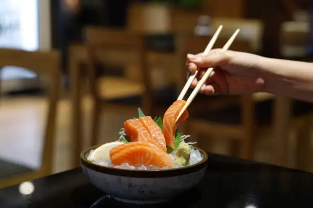 Photo of Hand holding salmon sashimi using chopsticks - Raw fresh salmon sliced served on ice with wasabi.