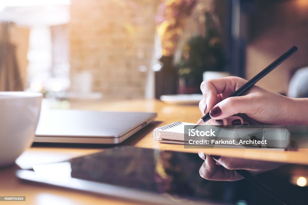 Closeup image of woman's hand writing on a blank notebook with laptop , tablet and coffee cup on wooden table background Pencil Stock Photo