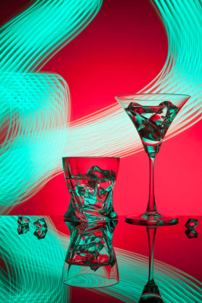 Multicolored glasses filled with alcoholic drinks, with splases of ice cubes falling inside, standing on the mirror surface. Black background. Conceptual, celebrated, commercial design. Closeup.