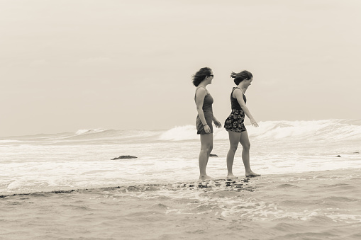 Teenager girls  beach holidays walking talking time on tidal pool wall with overcast ocean waves on horizon.
