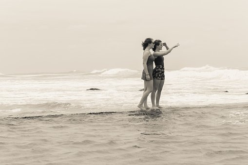 Teenager girls  beach holidays walking talking time on tidal pool wall with overcast ocean waves on horizon vintage cream boack and white photo.