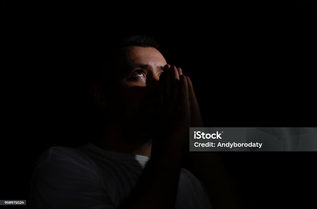 man praying to God in the dark room man praying to God in the dark room over concrete wall with window light effected, can be used for christian background or trust concept, copy space. prayer on a black background in a white t-shirt Adult Stock Photo