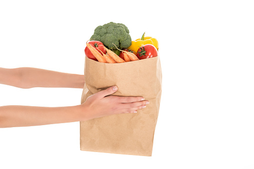 close-up partial view of woman holding paper bag with fruits and vegetables isolated on white