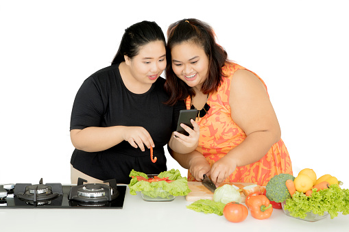 Image of two fat women looking at the smartphone while making healthy salad, isolated on white background