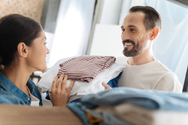 Jolly optimistic couple doing laundry Mutual help. Cheerful appealing merry couple helping each other with laundry while laughing and gazing at each other laundry husband housework men stock pictures, royalty-free photos & images