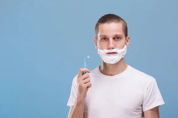 Photo of A guy, with foam for shaving on face, is holding a razor. Hygiene concept. Blue background.