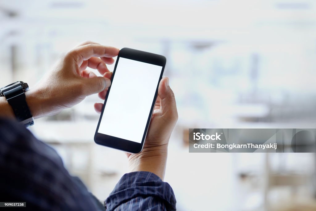 Mockup image of man hands holding black mobile phone with blank white screen in office. Computer Monitor Stock Photo