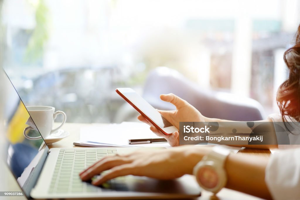 Young woman looking smartphone and using laptop computer while working in office. Mobile Phone Stock Photo