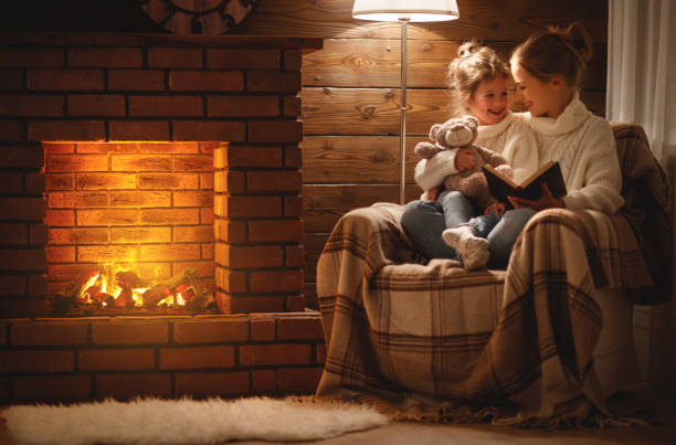 glückliche familie, mutter und kind tochter lesen buch über winterabend kamin - behaglich stock-fotos und bilder