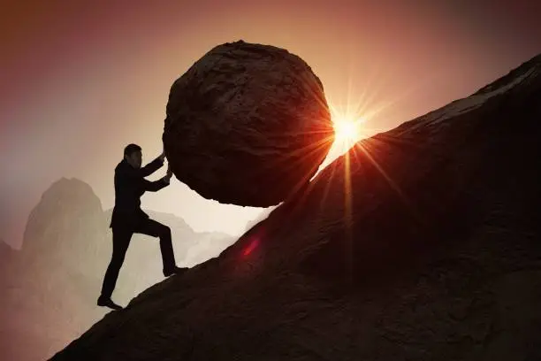 Photo of Sisyphus metaphore. Silhouette of businessman pushing heavy stone boulder up on hill.