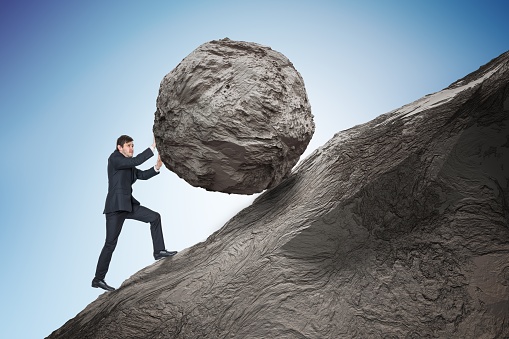 Sisyphus metaphore. Young businessman pushing heavy stone boulder up on hill.