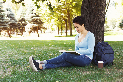 Young smiling woman reading book in park, preparing for exams at university or college, sitting on grass. Education concept, copy space