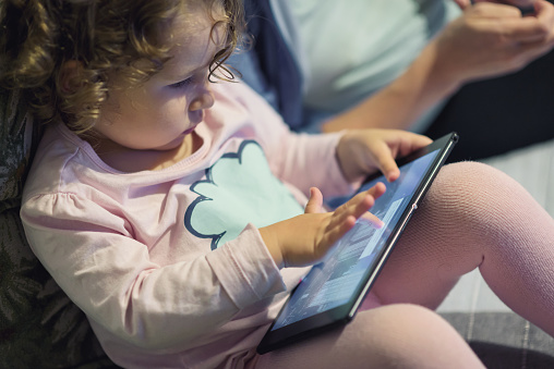Little girl sits near her mother and uses a digital tablet. Two-year child looks electronic device at home.