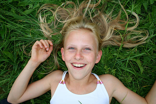 A young girl with long blonde hair lies on a green lawn, her head resting on her hand and a bunch of red flowers held close to her face. She smiles gently, her eyes reflecting a dreamy expression. The outdoor setting, with soft natural light and blurred trees in the background, emphasizes her serene mood and the beauty of the simple, tranquil moment.