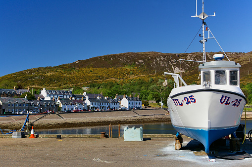Ullapool, Scotland - May 27th, 2012 - White fishing boat ashore on the pier with harbor, picturesque waterfront buildings and mountain range in the background
