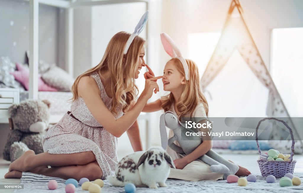 Mom and daughter preparing for Easter celebration Attractive young woman with little cute girl are preparing for Easter celebration. Mom and daughter wearing bunny ears are having fun with Easter bunny while sitting on the floor at home. Easter Stock Photo