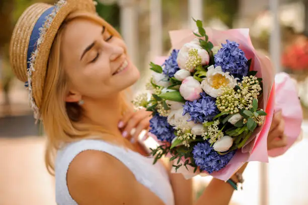 blonde pretty woman in white dress and straw hat holds a bouquet of hyacinths and peonies in pink wrapping-paper