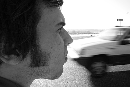 Profile of a young man.  SUV is seen passing on street in background.