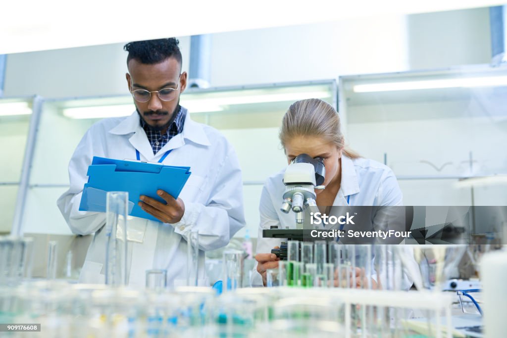 Scientists Doing Research in Lab Portrait of young Middle-eastern scientist taking notes on clipboard while working on medical research in laboratory while his female colleague looking in microscope Laboratory Stock Photo