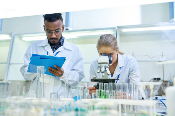 Scientists Doing Research in Lab Portrait of young Middle-eastern scientist taking notes on clipboard while working on medical research in laboratory while his female colleague looking in microscope clipboard photos stock pictures, royalty-free photos & images