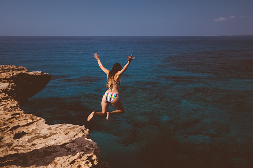 Young active woman jumping off cliff and falling into ocean on tropical island summer holidays